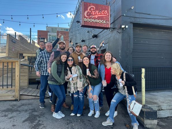 Group of friends posing outside an urban bar with string lights and a wooden patio, smiling and holding drinks on a sunny day