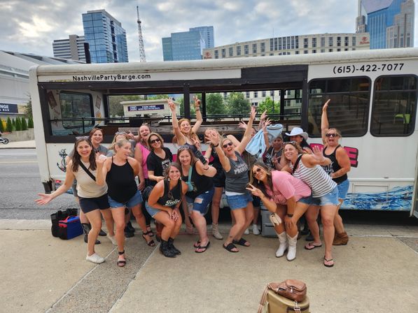 Energetic group of women posing and cheering beside a white party shuttle parked on a city street, downtown skyscrapers in the background, casual summer outfits and cowboy boots