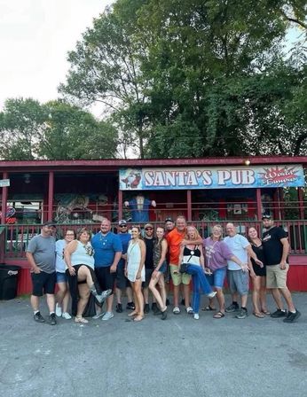 About 15 adults smiling and posing together outside a red roadside pub with a Santa-themed banner, tall trees and a casual summer vibe.