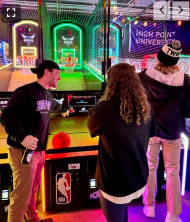 Three friends playing a neon-lit arcade basketball game in an indoor entertainment center with vibrant LED hoops and an NBA-branded cabinet.