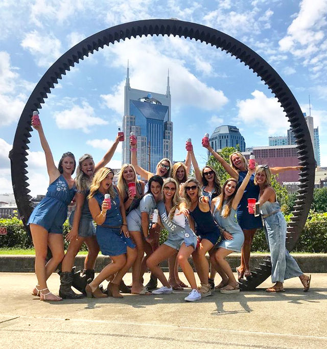 Energetic bachelorette crew in denim raising drinks and posing under a giant circular gear sculpture with Nashville downtown skyline behind them