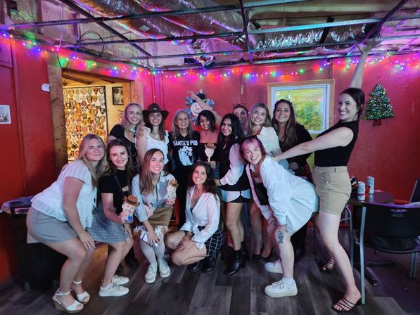 Group of smiling women posing for a festive indoor bar photo under colorful string lights and holiday decorations