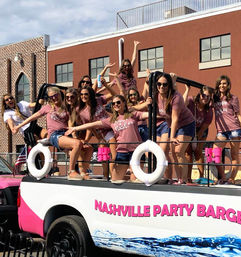 Group of women in matching shirts partying and posing on a pink-and-white party barge float in a sunny downtown Nashville street parade, brick buildings behind them.