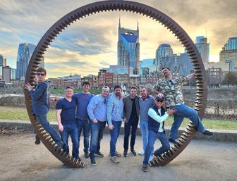 Group of friends posing inside a giant rusted gear sculpture on the Nashville riverfront with downtown skyline and twin‑spired skyscraper at sunset