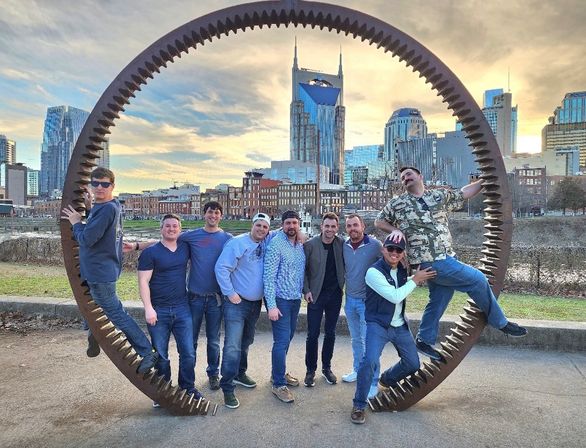 Group of friends posing inside a giant rusted gear sculpture on the Nashville riverfront with downtown skyline and twin‑spired skyscraper at sunset