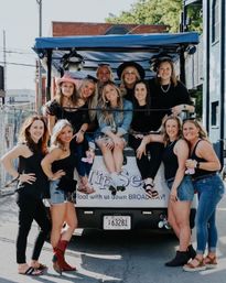 Group of smiling women posing on and around a covered party cart on a sunny downtown street near Broadway, wearing casual summer outfits, denim and cowboy boots for a fun girls' outing.