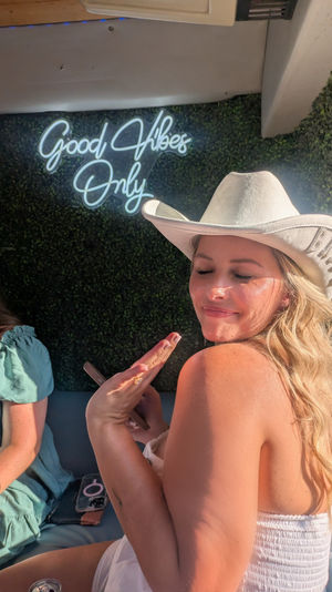 Sunlit woman in a white cowboy hat and strapless top smiling with eyes closed in front of a green hedge wall and neon sign reading 'Good Vibes Only' — casual party photo vibe