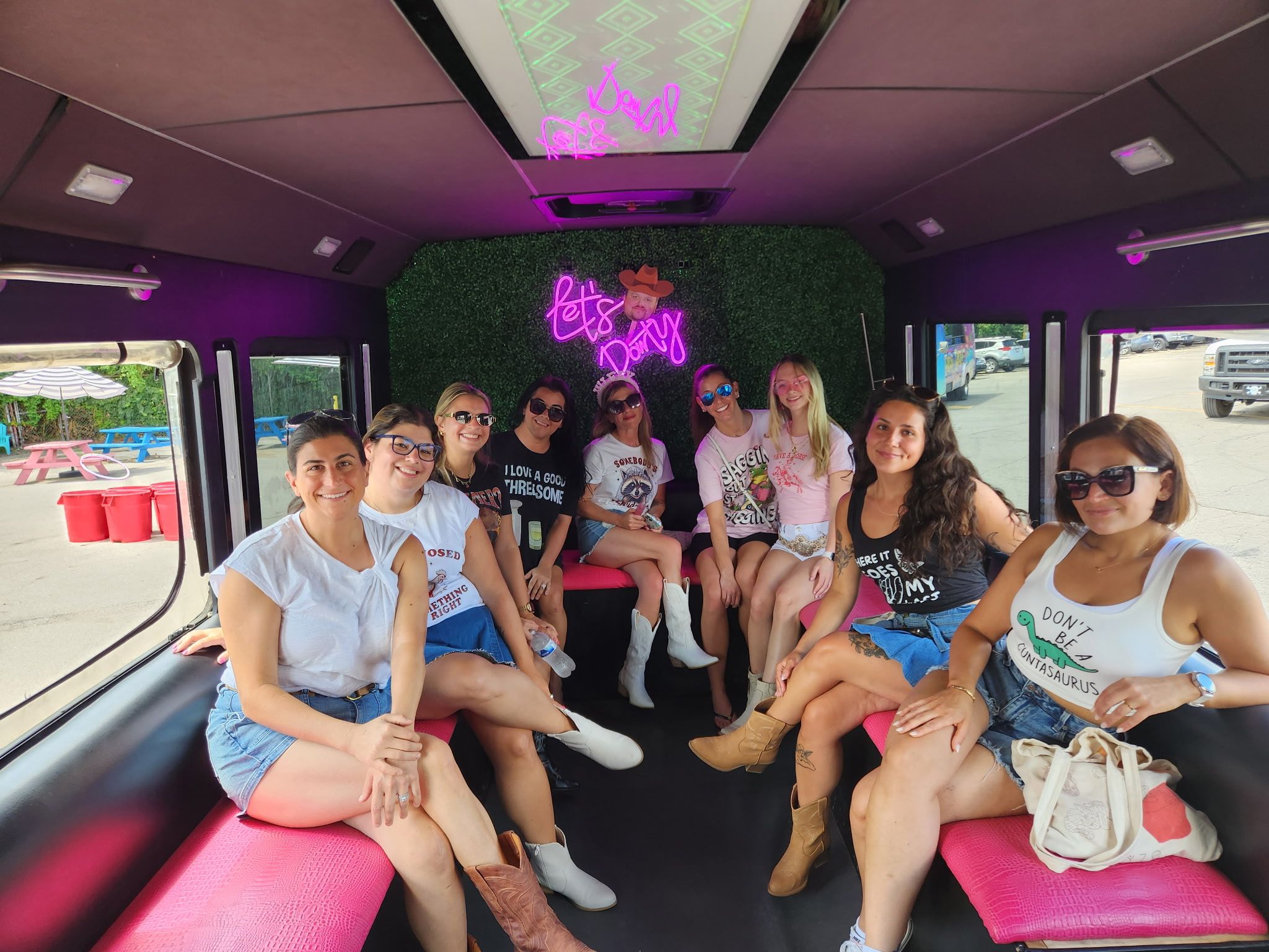 Group of friends inside a pink-seated party bus interior with a neon "Let's Party" sign, smiling in casual summer outfits and cowboy boots during a daytime girls' weekend/bachelorette ride