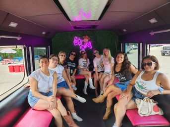 Group of friends inside a pink-seated party bus interior with a neon "Let's Party" sign, smiling in casual summer outfits and cowboy boots during a daytime girls' weekend/bachelorette ride