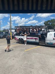 Group of women riding an open-air party bus in a sunny parking lot, a bride-to-be in a white dress, boots and hat posing in front while a friend photographs them under a bright blue sky with fluffy clouds.
