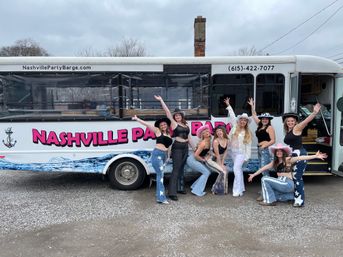 Smiling women in cowgirl hats and star-print jeans striking playful poses in front of a white party bus in Nashville, TN on a cloudy day