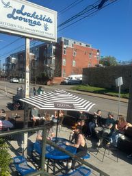 Sunny urban bar patio with people enjoying drinks under a striped umbrella at blue metal tables beside a street and red-brick apartment building, string lights overhead.