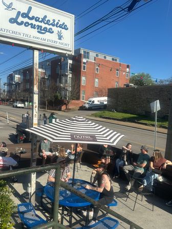 Sunny urban bar patio with people enjoying drinks under a striped umbrella at blue metal tables beside a street and red-brick apartment building, string lights overhead.