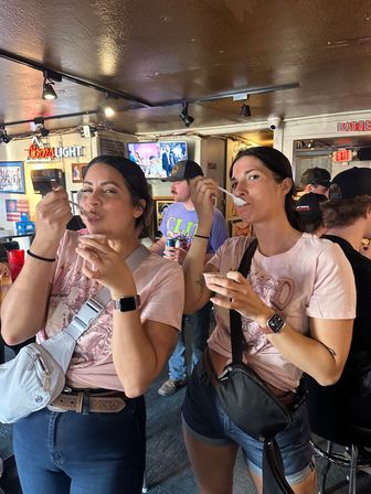 Two women in matching pink graphic tees and crossbody bags enjoying spoonfuls of dessert at a lively neighborhood bar, with TV screens, neon beer signage, and patrons in the background.