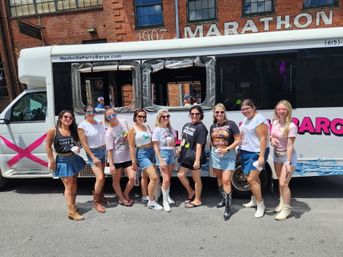 Group of women in summer outfits and cowboy boots posing by a white party bus with pink accents on a sunny downtown Nashville street in front of red brick warehouse buildings.