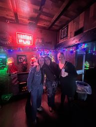 Three friends posing in a cozy holiday-decorated pub interior with colorful string lights, Santa decorations, a red neon sign and one holding a canned drink, festive nightlife scene.