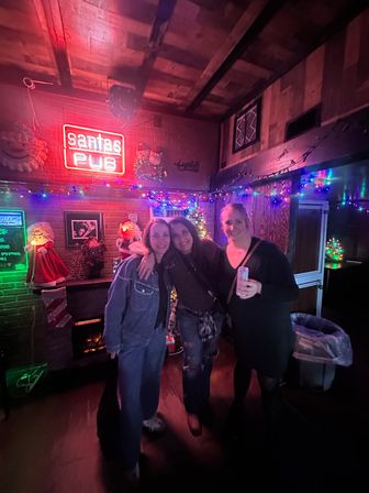 Three friends posing in a cozy holiday-decorated pub interior with colorful string lights, Santa decorations, a red neon sign and one holding a canned drink, festive nightlife scene.