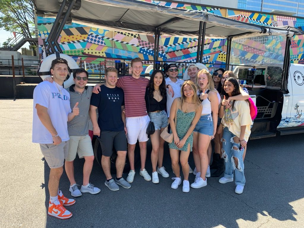 Group of friends posing in front of a colorful geometric mural beside an open-air party vehicle on a sunny urban street — casual summer outfits, sneakers, and big smiles.