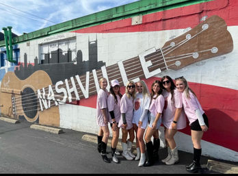 Eight friends in pink shirts and cowboy boots posing in front of a large painted guitar and Nashville skyline mural on an exterior wall