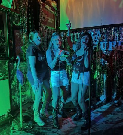 Three women singing karaoke on a tinsel-decorated bar stage with microphones, wearing denim shorts and cowboy boots under teal and purple neon lights, lively nightlife scene