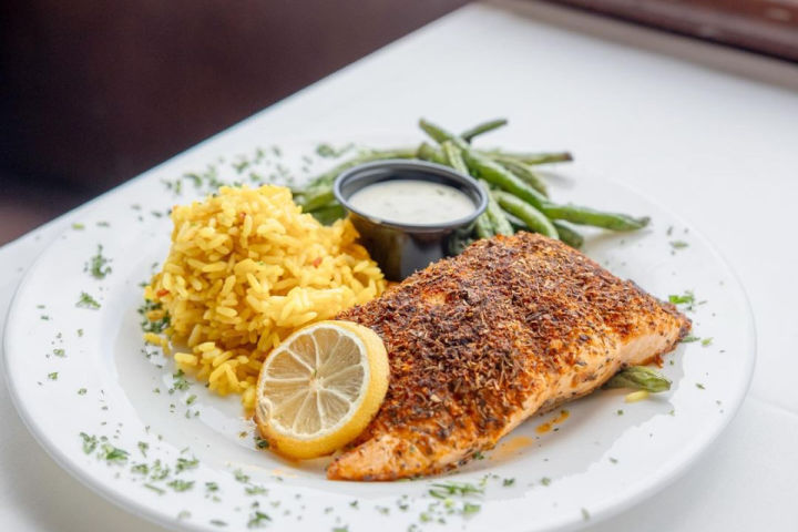 Grilled seasoned salmon fillet with lemon slice, yellow rice, green beans and creamy sauce on a white restaurant plate, garnished with chopped herbs