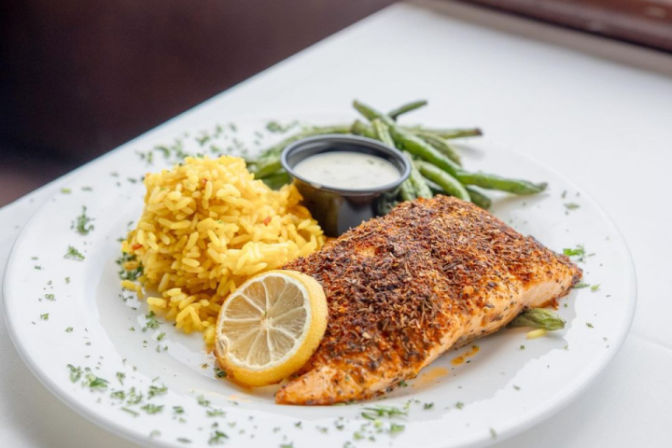 Grilled seasoned salmon fillet with lemon slice, yellow rice, green beans and creamy sauce on a white restaurant plate, garnished with chopped herbs