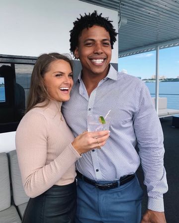 Smiling couple on a waterfront boat deck with city skyline in the background, woman holding a lime-garnished cocktail and both dressed in smart-casual attire.