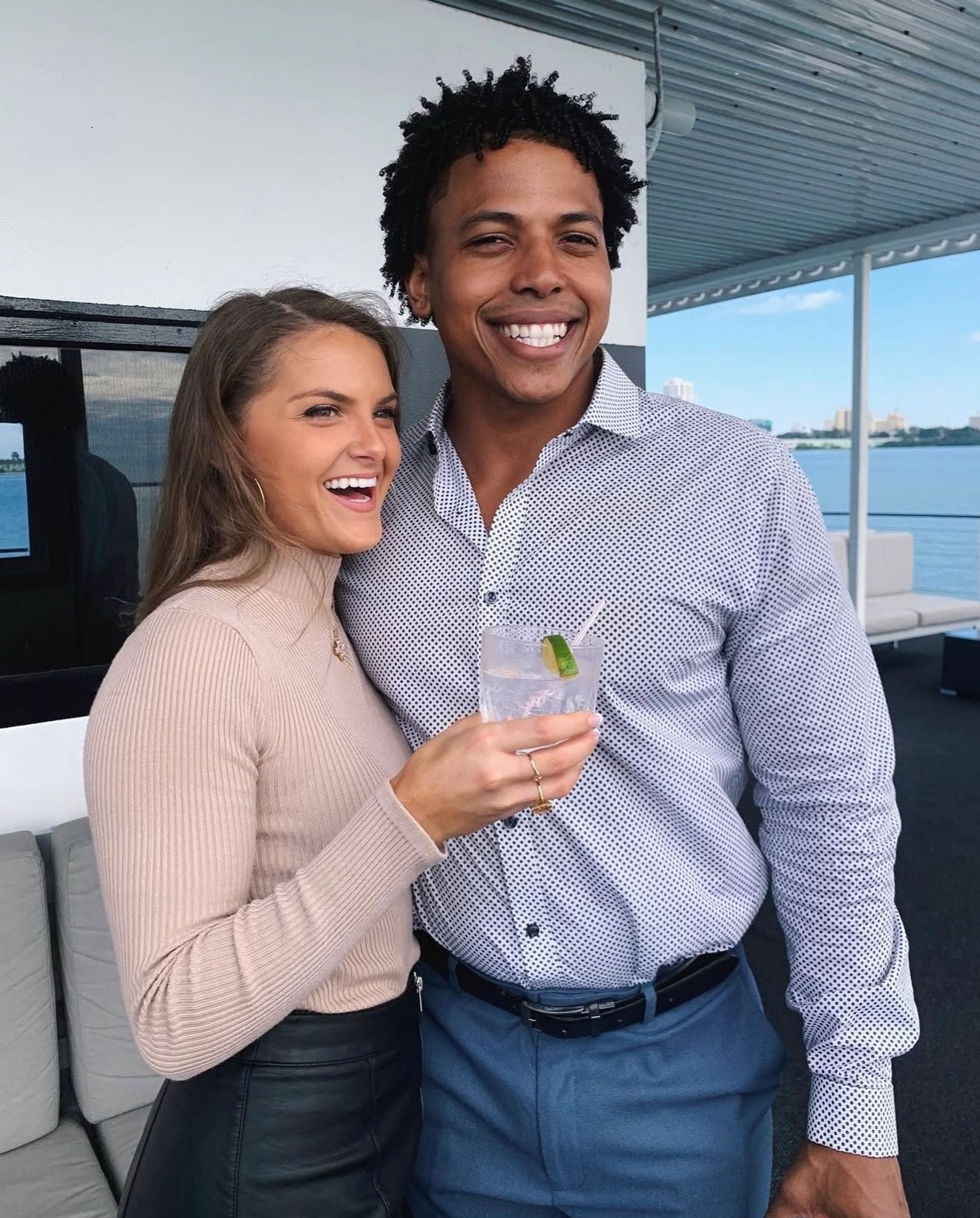 Smiling couple on a waterfront boat deck with city skyline in the background, woman holding a lime-garnished cocktail and both dressed in smart-casual attire.