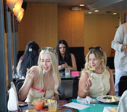 Two women enjoy brunch at a colorful, tropical‑themed cafe — fruit plates and cocktails on a painted table, warm wall lights and other diners in the background.