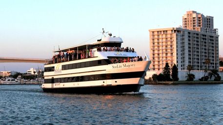 Sightseeing passenger yacht crowded with people cruising a coastal harbor at sunset past high-rise waterfront condominiums and a low bridge.