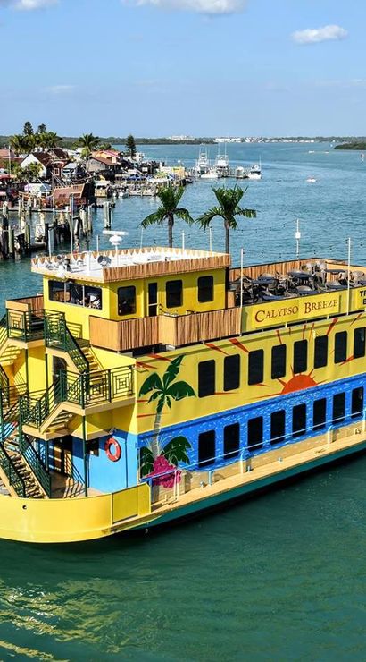 Colorful yellow double-decker tropical-themed party boat with palm murals and rooftop seating cruising past a busy seaside harbor under a sunny blue sky