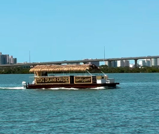 Tiki-style tour boat with a thatched roof cruising across a blue bay past a bridge and city skyline