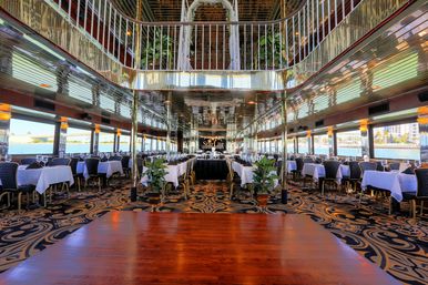 Elegant waterfront dinner cruise dining room aboard a yacht-style riverboat with white‑clothed tables, mirrored ceiling, patterned carpet and panoramic water views.