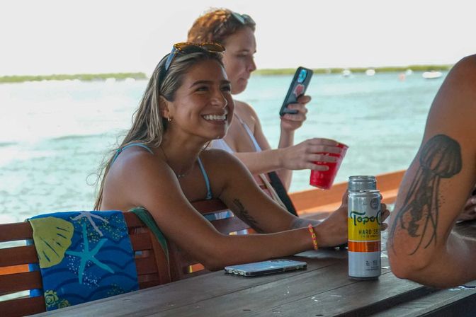 Smiling woman in a swimsuit on a sunny boat near turquoise water, friends passing drinks and checking a phone