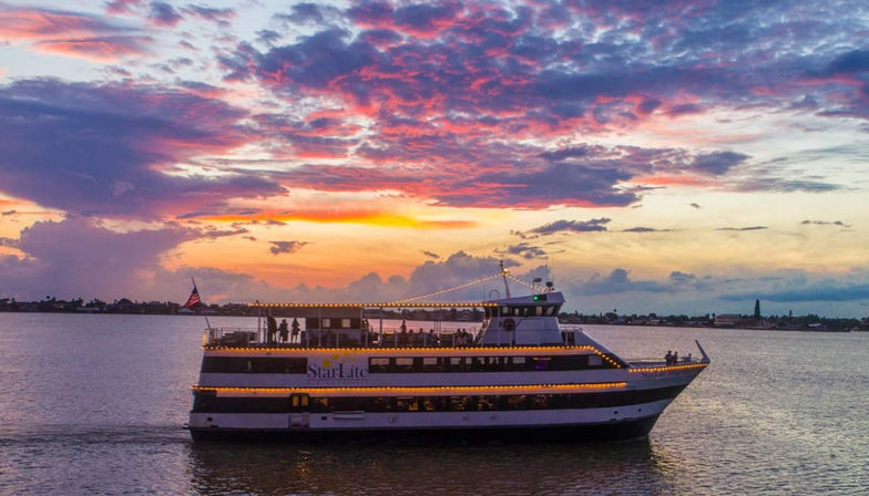 Lit-up dinner yacht cruising a calm coastal bay at sunset beneath vibrant pink and purple clouds