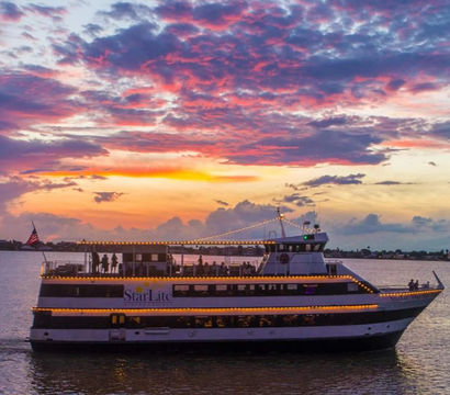 Lit-up dinner yacht cruising a calm coastal bay at sunset beneath vibrant pink and purple clouds