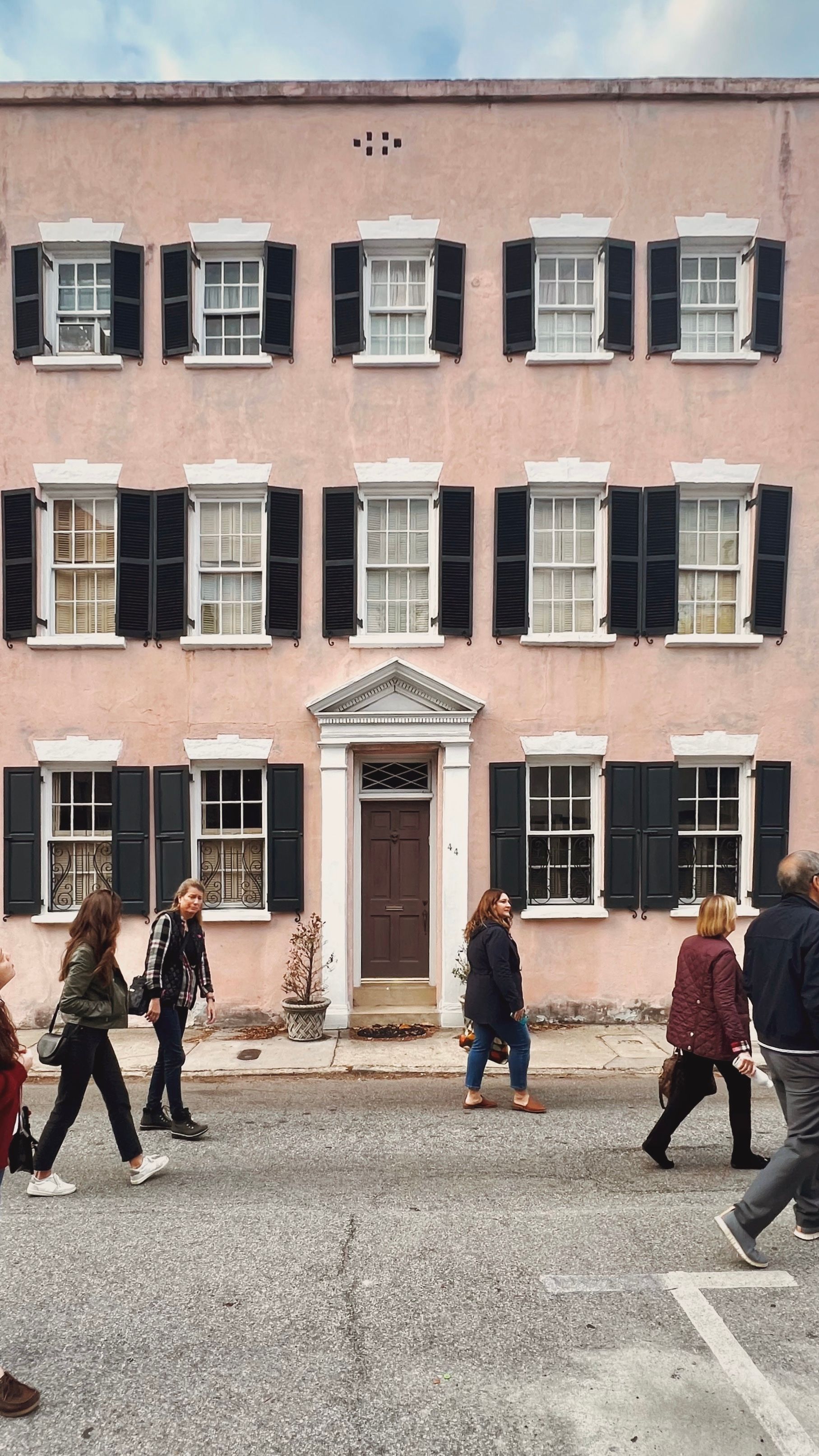 Three-story pastel-pink townhouse with white trim and black shutters, central brown pedimented door, potted plants at the stoop and pedestrians strolling along the urban street
