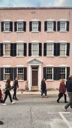 Three-story pastel-pink townhouse with white trim and black shutters, central brown pedimented door, potted plants at the stoop and pedestrians strolling along the urban street