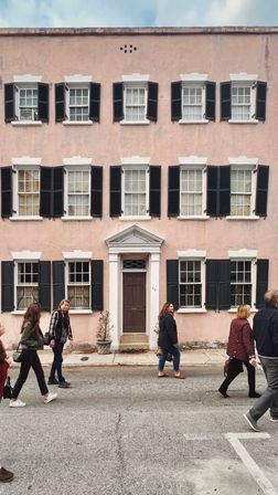 Three-story pastel-pink townhouse with white trim and black shutters, central brown pedimented door, potted plants at the stoop and pedestrians strolling along the urban street