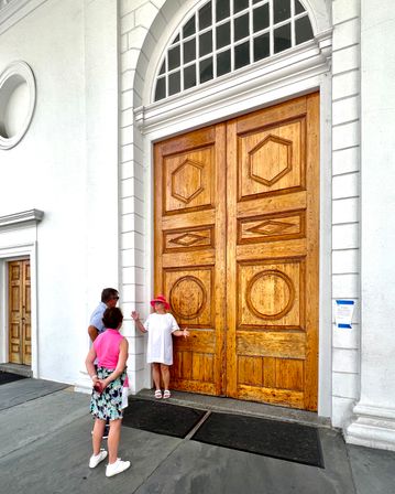 Three people on the sidewalk beside towering carved wooden double doors with an arched transom in a white neoclassical facade; a woman in a pink hat and white dress poses with her arms out.