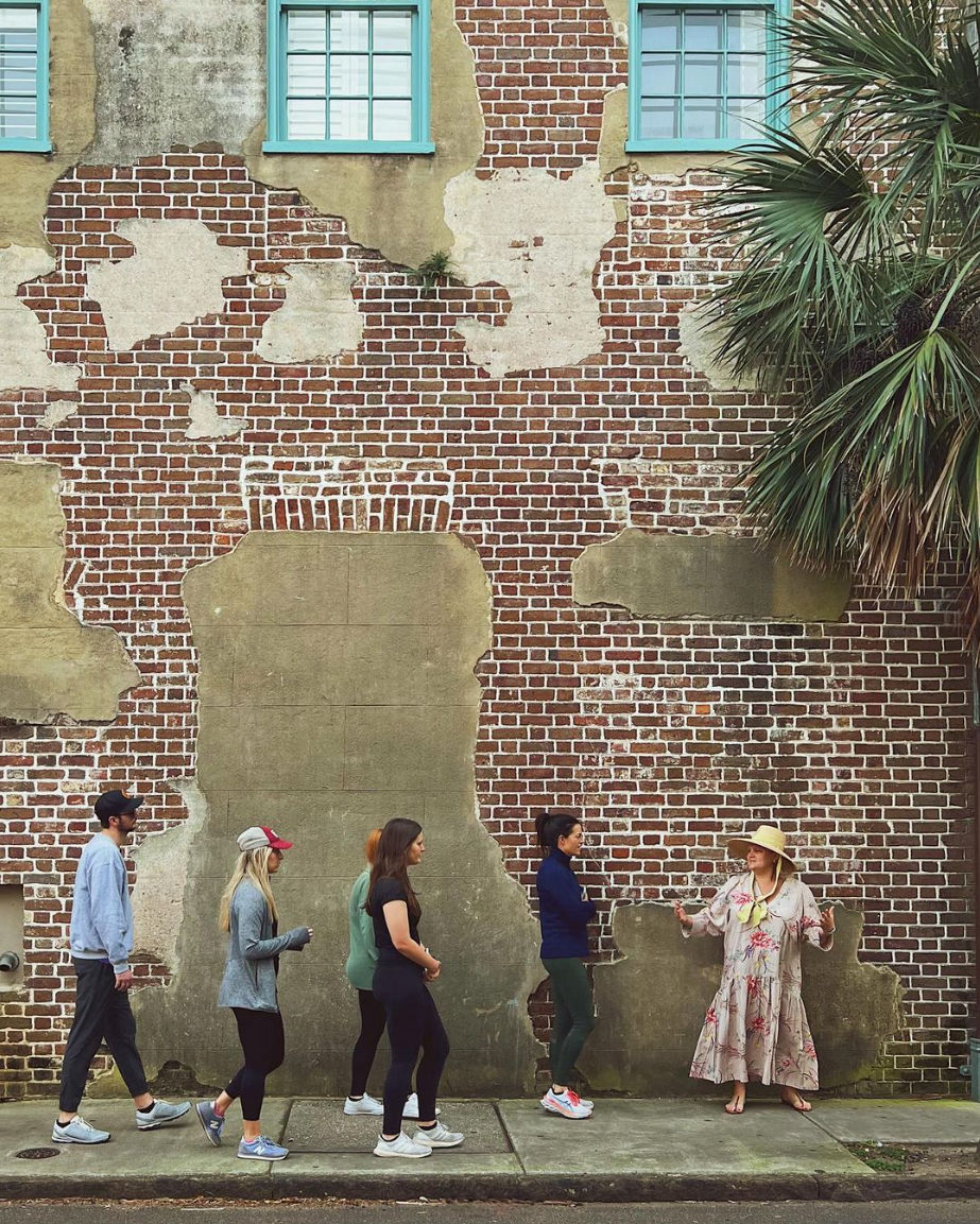 Walking group of five listening to a gesturing woman in a floral dress and sun hat in front of a weathered red brick wall with peeling plaster, turquoise windows, and a palm tree.
