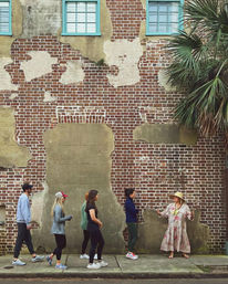 Walking group of five listening to a gesturing woman in a floral dress and sun hat in front of a weathered red brick wall with peeling plaster, turquoise windows, and a palm tree.