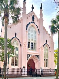 Pink Gothic Revival church facade with tall arched windows, decorative black iron trim and fence, flanked by palm trees on a sunny urban sidewalk