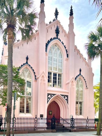 Pink Gothic Revival church facade with tall arched windows, decorative black iron trim and fence, flanked by palm trees on a sunny urban sidewalk