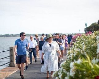 Sunny waterfront promenade with people strolling past blooming white flowers; a woman in a sunhat and a man with coffee among the crowd.