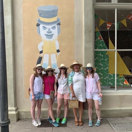 Five friends in summer outfits and straw hats smiling on a downtown sidewalk in front of a painted toy-character mural and festive window display.