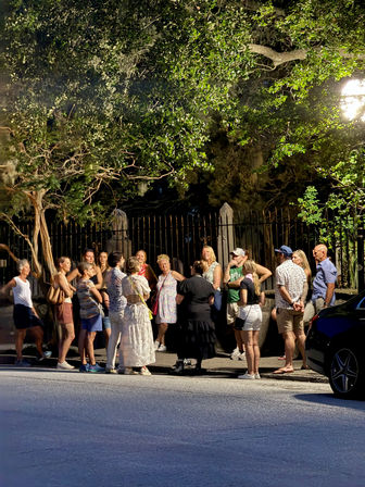 Group of casually dressed people gathered on a downtown city sidewalk at night beneath a large leafy tree and bright streetlamp, standing in front of an iron fence.