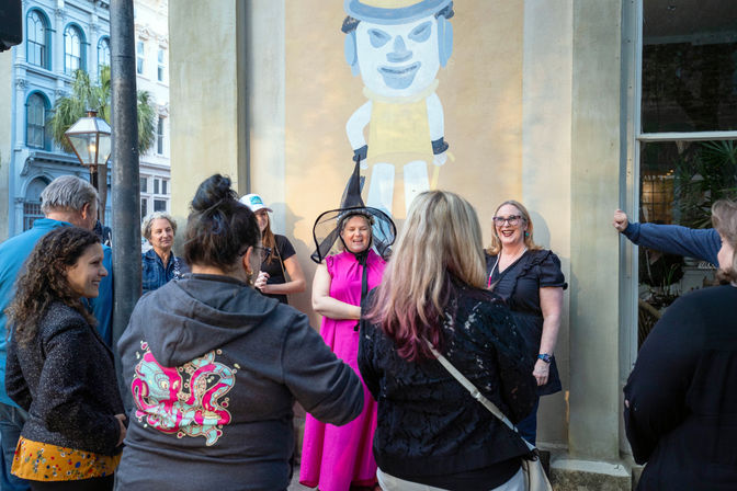Group gathered on a historic downtown street in front of a large mural, people laughing as a woman in a bright pink dress and witch hat poses