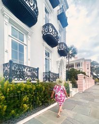 Person in a sunhat and pink floral dress strolling past pastel historic row houses with ornate black wrought-iron balconies and a palm tree on a Charleston sidewalk