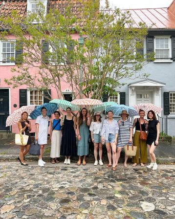 Group of women holding colorful patterned umbrellas on a cobblestone street in front of pastel pink and blue row houses beneath a leafy tree